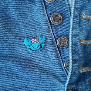 May include: A blue crab pin with pink accents on a denim background. The pin is attached to a pair of jeans with silver buttons.