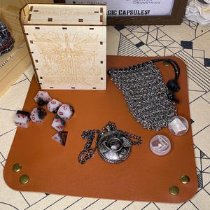 May include: A brown leather gaming mat with a metal chainmail bag, a silver pendant with wings and a dice tower with a wood grain finish. The dice tower has a design of a sword with wings on the front. The mat has two brass grommets on the left and two on the right. The mat also has a silver dice with red numbers and a clear glass dice.