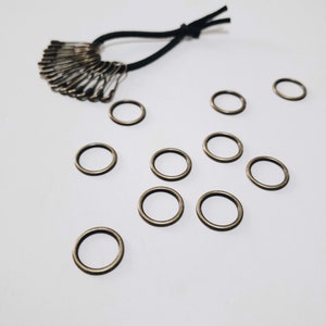 May include: A selection of small, bronze-coloured metal rings and a cluster of safety pins attached to a black elastic band. The rings are scattered on a white surface, with the safety pins arranged in a fan shape.
