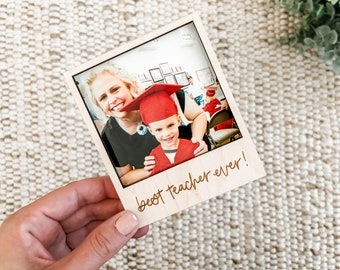Puede incluir: Un marco de fotos de madera con una foto de una maestra y un niño con un gorro de graduación. El marco tiene el texto "¡la mejor maestra de la historia!" escrito en él.