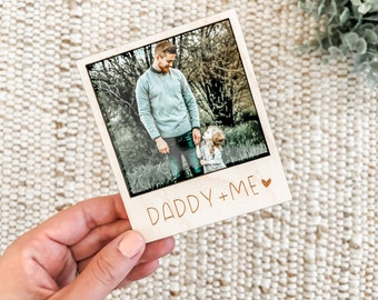Pode incluir: Um porta-retratos de madeira com uma foto de um pai e uma filha de mãos dadas. O porta-retratos tem um fundo branco e o texto "Daddy + Me" com um símbolo de coração.