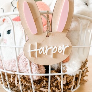 May include: A wooden bunny-shaped Easter basket tag with pink ears and the name "Harper" written in white. The tag is hanging from a white wire basket filled with brown shredded paper and stuffed animals.