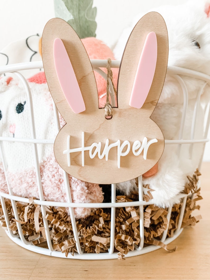 May include: A wooden bunny-shaped Easter basket tag with pink ears and the name "Harper" written in white. The tag is hanging from a white wire basket filled with brown shredded paper and stuffed animals.