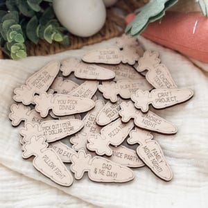 May include: A collection of wooden carrot-shaped tokens with various activities and phrases engraved on them. The tokens include phrases like "Picnic in the Park," "Family Movie Night," and "Dad & Me Day!" The tokens are light brown with dark brown edges.
