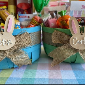 May include: Two blue and green Easter baskets filled with candy, bath bombs, and other treats. The baskets are decorated with burlap bows and bunny-shaped wooden tags that say "Penn" and "Carly".