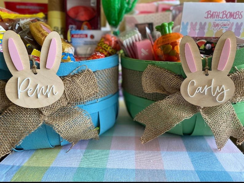 May include: Two blue and green Easter baskets filled with candy, bath bombs, and other treats. The baskets are decorated with burlap bows and bunny-shaped wooden tags that say "Penn" and "Carly".