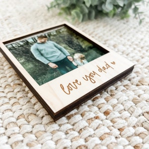 May include: A wooden photo frame with a picture of a father and child in a forest. The frame has a brown border and the words "love you dad" are engraved on the bottom.