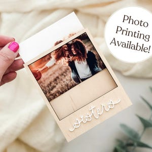 May include: A wooden photo frame with a picture of two people. The word "sisters" is written in cursive on the frame. The text "Photo Printing Available!" is in a white circle.
