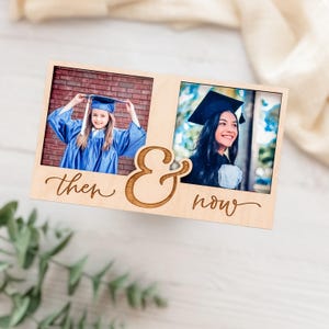May include: A wooden photo frame with two openings, each holding a graduation photo. The frame is engraved with the words "then & now" in a cursive font. The photos show a young person in a blue graduation gown.