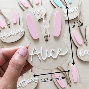 May include: Wooden bunny-shaped ornaments with pink ears and white names. The ornaments are 3.75 inches tall and 2.63 inches wide. The names on the ornaments are Hadley, Alice, and Matthias.