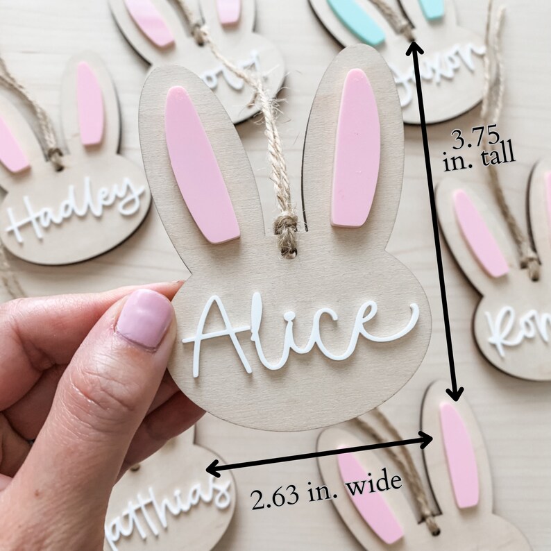 May include: Wooden bunny-shaped ornaments with pink ears and white names. The ornaments are 3.75 inches tall and 2.63 inches wide. The names on the ornaments are Hadley, Alice, and Matthias.