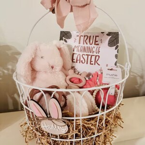 May include: A white wire basket filled with baby items for Easter. The basket is decorated with a pink bow and a sign that says "True Meaning of Easter". The basket contains a pink bunny plush toy, a wooden teether, a pink and white headband, a pink and white swaddle, and a wooden sign that says "Happy".