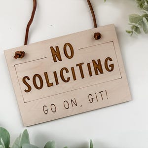 May include: A wooden sign with the text "NO SOLICITING GO ON, GIT!" in brown lettering. The sign is hanging from a brown leather cord.
