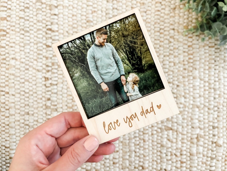 May include: A wooden photo frame with a picture of a father and daughter in a forest. The frame has a light brown wood finish and the text "love you dad" is engraved on the bottom.
