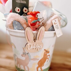 May include: A white bucket with a unicorn and rainbow design. The bucket is filled with a variety of items, including a plush bunny ear headband, a troll doll, and a wooden bunny ear tag with the name "Oakley" written on it.