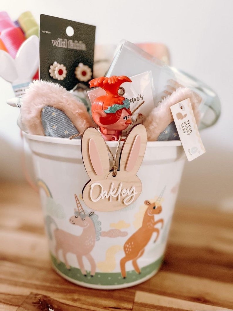 May include: A white bucket with a unicorn and rainbow design. The bucket is filled with a variety of items, including a plush bunny ear headband, a troll doll, and a wooden bunny ear tag with the name "Oakley" written on it.