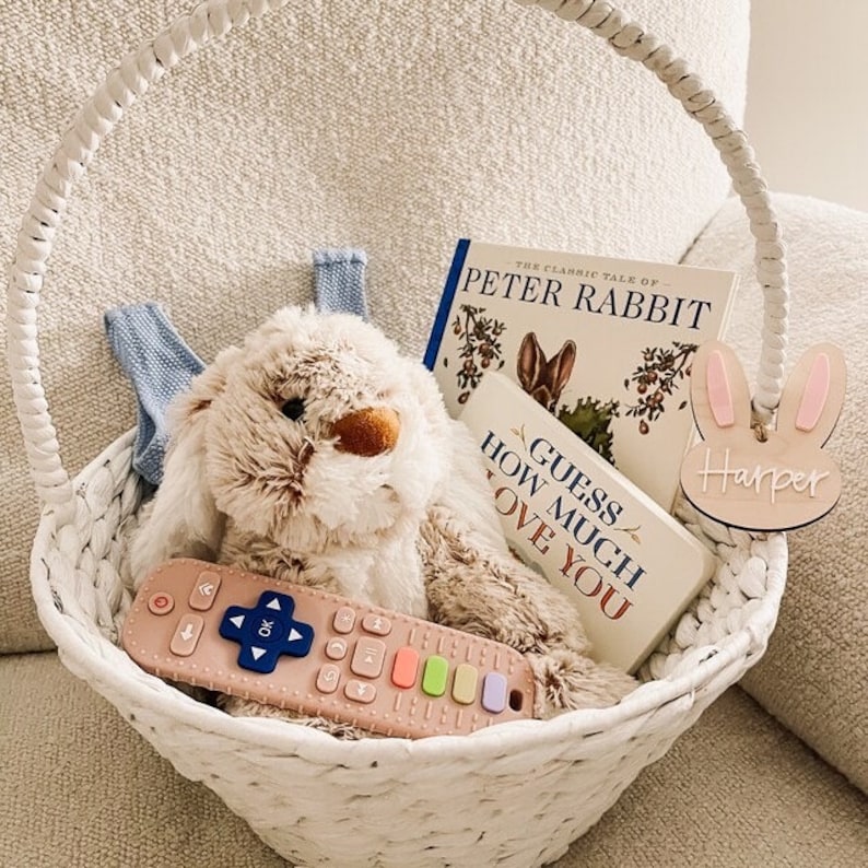May include: A white wicker basket filled with Easter gifts, including a plush bunny, a book titled "The Classic Tale of Peter Rabbit", a book titled "Guess How Much I Love You", and a wooden bunny ear tag with the name "Harper" on it.