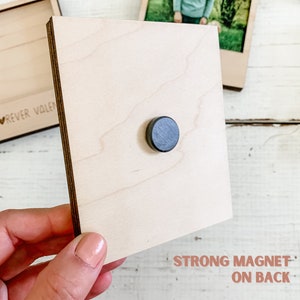 May include: A wooden photo frame with a strong magnet on the back. The frame is light brown and has a smooth surface. The magnet is black and round.