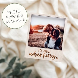 May include: A wooden photo frame in the style of a Polaroid picture, with the words "TO NEW adventures" engraved at the bottom. The frame holds a photo of two people. A circular graphic reads "PHOTO PRINTING AVAILABLE".