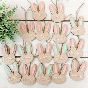 May include: Wooden bunny-shaped tags with pastel pink and blue ears and names written in white. The names include Kaiyla, Joelle, Magnus, Josephine, Lennon, Athena, Wrenley, Danni, August, McCartney, Duke, Dawson, Harper, James, Faith, and Scarlett.