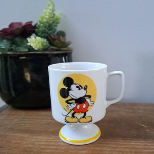 May include: White ceramic mug with a yellow circle featuring a cartoon image of Mickey Mouse. Mickey is wearing red shorts and yellow shoes. The mug has a white pedestal base with a yellow stripe around the top.