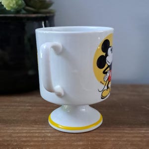 May include: White ceramic mug with a yellow and black Mickey Mouse design. The mug has a pedestal base and a handle.
