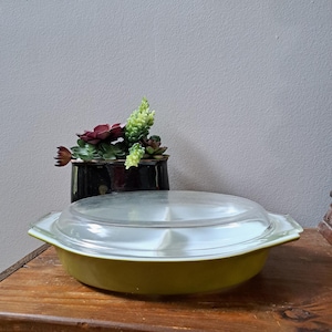 May include: A vintage green Pyrex casserole dish with a clear glass lid. The dish has a divided interior and is perfect for serving multiple dishes.