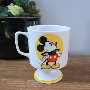 May include: White ceramic mug with a yellow circle and a cartoon image of Mickey Mouse in red shorts and yellow shoes. The mug has a yellow band around the bottom and a white pedestal base.