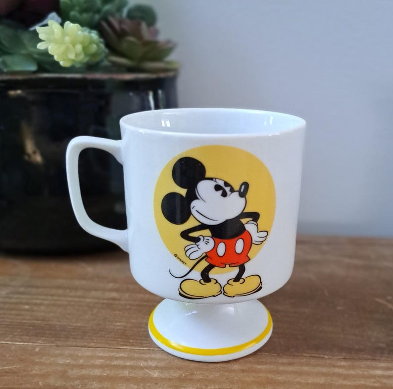 May include: White ceramic mug with a yellow circle and a cartoon image of Mickey Mouse in red shorts and yellow shoes. The mug has a yellow band around the bottom and a white pedestal base.