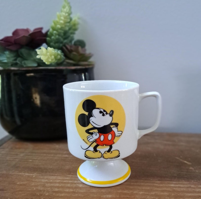 May include: White ceramic mug with a yellow circle featuring a cartoon image of Mickey Mouse. Mickey is wearing red shorts and yellow shoes. The mug has a white pedestal base with a yellow stripe around the top.