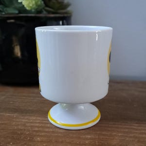 May include: White ceramic mug with a yellow and black design. The mug has a pedestal base.