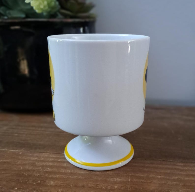 May include: White ceramic mug with a yellow and black design. The mug has a pedestal base.