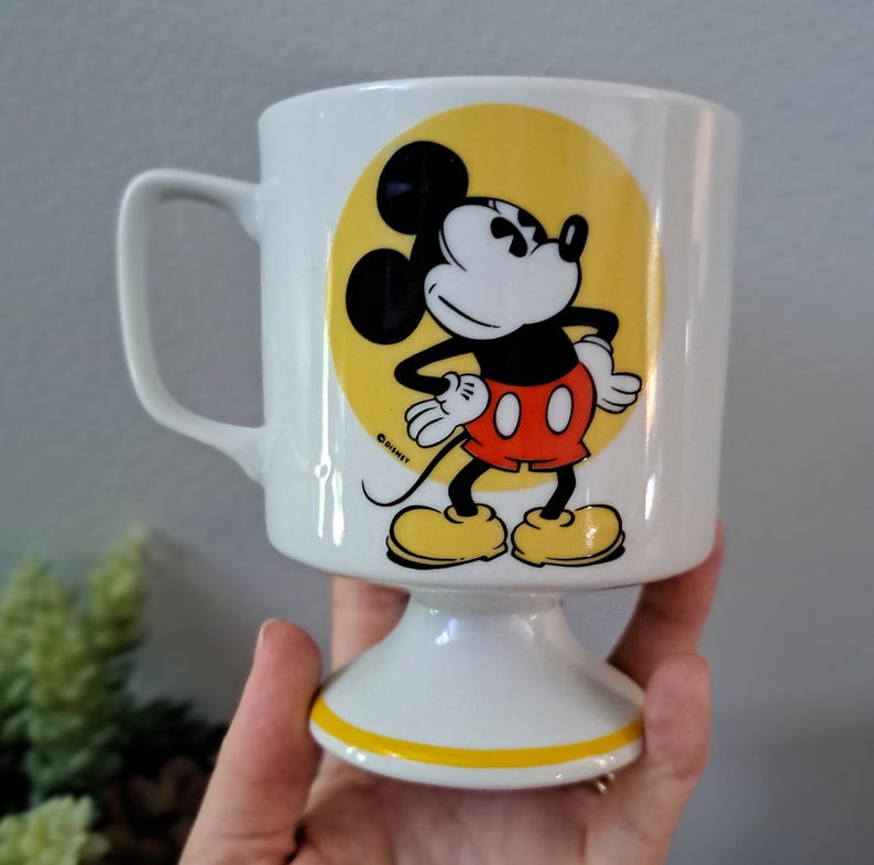 May include: White ceramic mug with a yellow circle featuring a cartoon image of Mickey Mouse. Mickey is wearing his signature red shorts and yellow shoes. The mug has a white pedestal base with a yellow stripe.