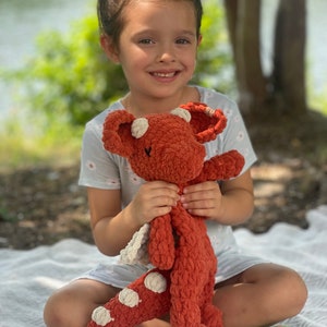 May include: A handmade, rust-colored crochet dragon plush toy with cream-colored accents. The dragon has large ears, a long body, and white spots. The plush is soft and cuddly, perfect for a child's playtime or as a decorative item.