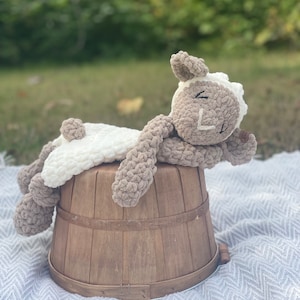 May include: A crocheted plush lamb toy with brown and white fur, lying on a wooden barrel. The lamb is resting its head on the barrel and has a sweet expression on its face.