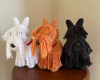 May include: Three crocheted stuffed dogs, one white, one orange, and one black, all with long shaggy hair. The dogs are sitting on a wooden surface.