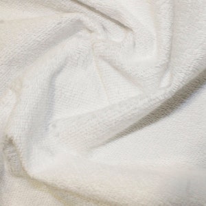May include: Close-up of a white, textured fabric with a soft, plush appearance. The material is folded and draped, creating shadows and highlights that emphasize its texture. The fabric appears to be a thick knit or terry cloth, suggesting comfort and warmth.