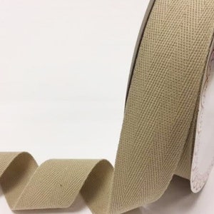 May include: A roll of beige herringbone ribbon. The ribbon is unravelling, displaying its textured weave and pattern. The spool is partly visible, with a decorative edge.