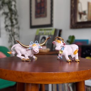 May include: Two porcelain cow figurines in a pale pink finish. The left cow features long gold-toned horns and a blue bow with small flowers. The right cow has a gold-toned ring on its head and a garland of colorful flowers around its neck.