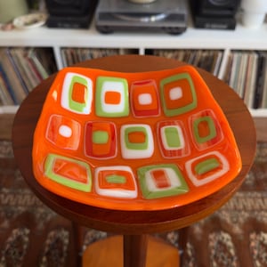 May include: A square fused glass dish featuring a vibrant orange base with a repeating pattern of nested rectangular shapes in white, lime green, and bright orange. The glass has a smooth, glossy finish with slightly curved, raised edges.