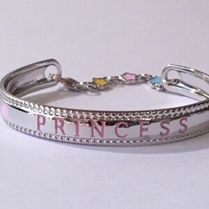 Pu&ograve; includere: Un braccialetto rigido in argento con un cuore rosa e la parola "PRINCESS" incisa in rosa. Il braccialetto ha una catena con tre piccole stelle in smalto giallo, rosa e blu.