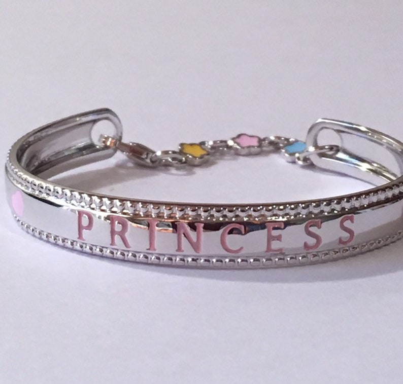 Pu&ograve; includere: Un braccialetto rigido in argento con un cuore rosa e la parola "PRINCESS" incisa in rosa. Il braccialetto ha una catena con tre piccole stelle in smalto giallo, rosa e blu.