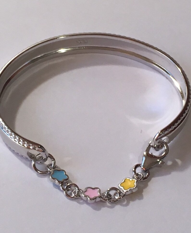 Pu&ograve; includere: Un braccialetto rigido in argento con una catena di quattro ciondoli smaltati. I ciondoli hanno la forma di stelle e sono di colore blu, rosa e giallo.