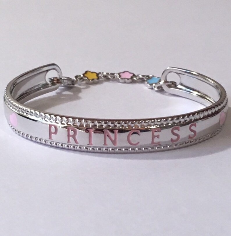 Pu&ograve; includere: Un braccialetto rigido in argento con un'iscrizione in smalto rosa che recita "PRINCESS". Il braccialetto ha una piccola catena con tre stelle in smalto giallo, rosa e blu.