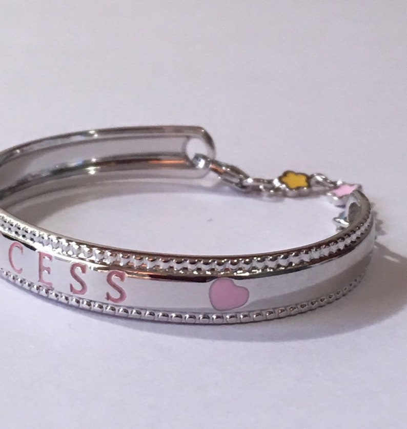 Pu&ograve; includere: Un braccialetto d'argento con un ciondolo a forma di cuore rosa e la parola "SUCCESS" incisa sulla fascia. Il braccialetto ha una catena delicata con piccoli ciondoli gialli e rosa.
