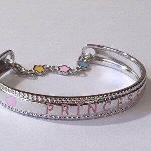 Pu&ograve; includere: Un braccialetto rigido in argento con un'iscrizione in smalto rosa che recita "PRINCESS". Il braccialetto ha un piccolo ciondolo a forma di cuore e una catena con tre stelle in smalto giallo, rosa e blu.