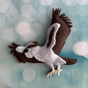 May include: A felt bird in flight, featuring brown and white coloring. The bird has outstretched wings and detailed features, set against a soft blue and white background. The bird's design is simple and charming.