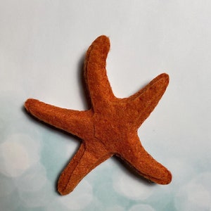 May include: A handcrafted orange felt starfish with five distinct points. The felt has a textured appearance. The starfish is set against a light blue and white bokeh background, evoking a beach or ocean theme.