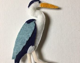 May include: A felt heron figurine in shades of white, light blue, and navy blue. The bird has a long yellow beak and stands on two cream-colored legs. The heron is a handmade craft item.
