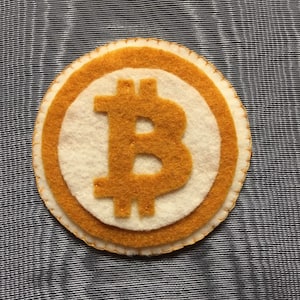 May include: A handmade felt Bitcoin coin with a gold-colored Bitcoin symbol on a white background, surrounded by a gold border. The coin is stitched with gold thread and rests on a striped fabric.
